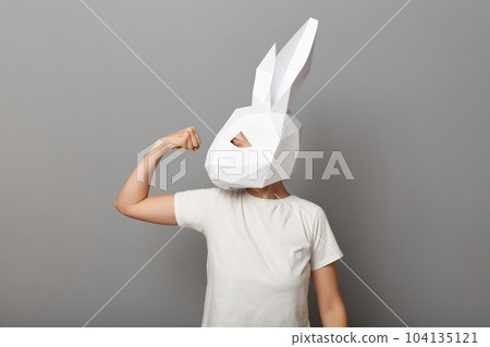 Image of strong unrecognizable woman wearing white t shirt and paper rabbit mask standing isolated over gray background, raised her arm, showing biceps and her power. Image of strong unrecognizable woman wearing white t shirt and paper rabbit mask standing isolated over gray background, raised her arm, showing biceps and her power. 104135121