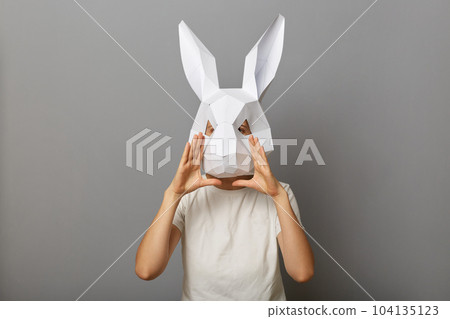 Photo of unknown woman wearing white T-shirt and paper rabbit mask, keeps hands near mouth, screaming loud, making announcement, telling advertisement, standing isolated over gray background. Photo of unknown woman wearing white T-shirt and paper rabbit mask, keeps hands near mouth, screaming loud, making announcement, telling advertisement, standing isolated over gray background. 104135123