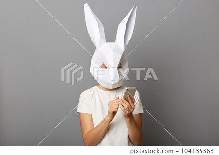 Indoor shot of female wearing bunny mask and orange sweatshirt posing isolated over gray background, holding smart phone in hands, using mobile phone, browsing internet. 104135363