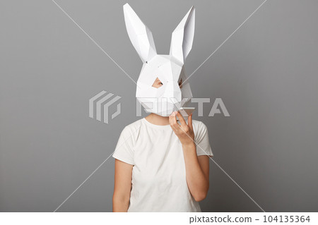 Indoor shot of anonymous woman wearing white t shirt and paper rabbit mask holding mobile phone recording voice message standing isolated over gray background. 104135364