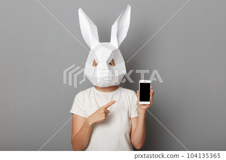 Portrait of unrecognizable woman wearing white t shirt and paper rabbit mask standing isolated over gray background, showing smart phone with empty screen, pointing at copy space. 104135365