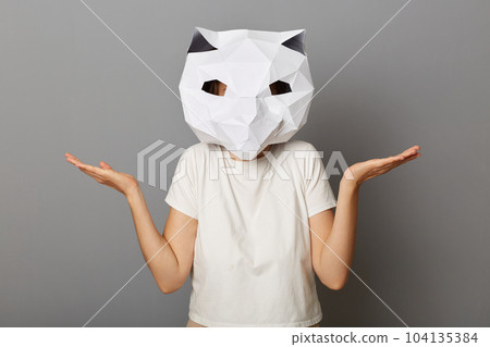 Portrait of uncertain helpless unknown woman wearing cat paper mask and white T-shirt posing isolated over gray background, shrugging shoulders, spread hands, doesn't know answer. 104135384