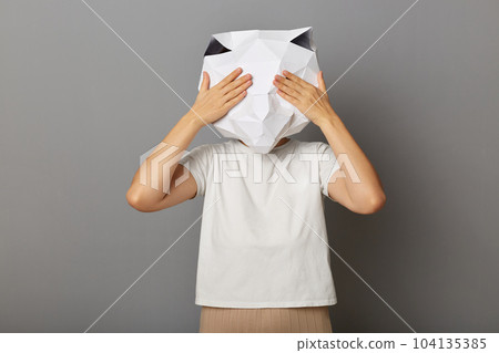 Indoor shot of unknown female wearing cat mask and white t-shirt isolated over gray background, standing covering her eyes with palms, hiding her face with hands. Indoor shot of unknown female wearing cat mask and white t-shirt isolated over gray background, standing covering her eyes with palms, hiding her face with hands. 104135385