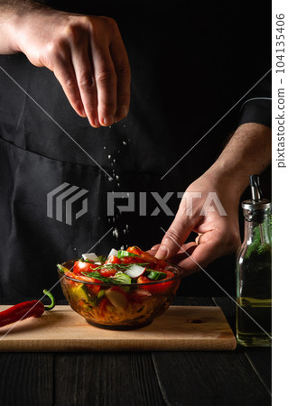 The chef cook sprinkles salted fresh vegetable salad in plate on a wooden table. Cooking healthy food in the kitchen in a restaurant The chef cook sprinkles salted fresh vegetable salad in plate on a wooden table. Cooking healthy food in the kitchen in a restaurant 104135406