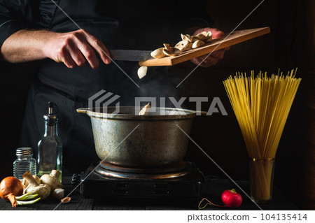 Chef prepares soup on the background of vegetables and spaghetti. Close-up of a cook hand pouring mushrooms into saucepan in a restaurant kitchen 104135414