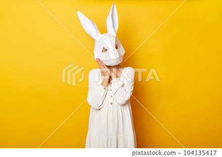 Portrait of sleepy woman wearing white dress and paper rabbit mask standing isolated over yellow background, needs to have nap, keeps palms near her face, drowsing. 104135417