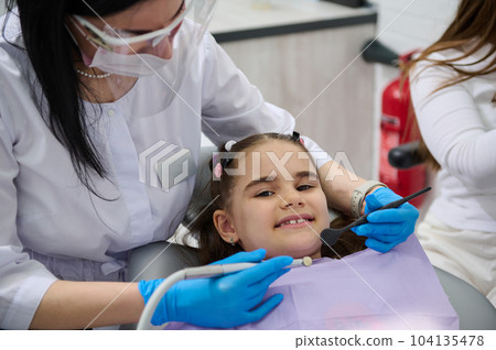 Smiling kid girl visiting dentist, curing tooth, putting braces in modern pediatric dentistry clinic. Female dentist, dental hygienist treating caries to little child patient. Oral health and hygiene 104135478