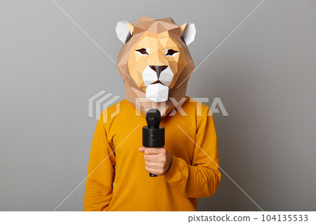 Indoor shot of anonymous man wearing casual sweater and paper king mask standing isolated over gray background, standing with microphone, reporter working, journalist, interview. 104135533