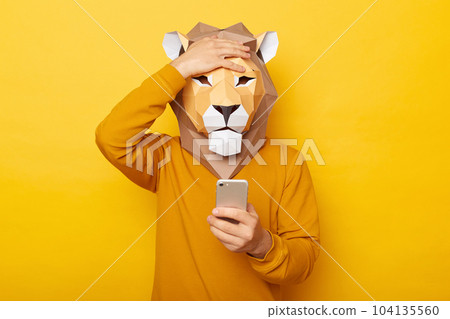 Photo of sad upset male wearing lion mask and orange sweatshirt posing isolated over yellow background, using mobile phone, having problems with internet connection, showing facepalm gesture. Photo of sad upset male wearing lion mask and orange sweatshirt posing isolated over yellow background, using mobile phone, having problems with internet connection, showing facepalm gesture. 104135560