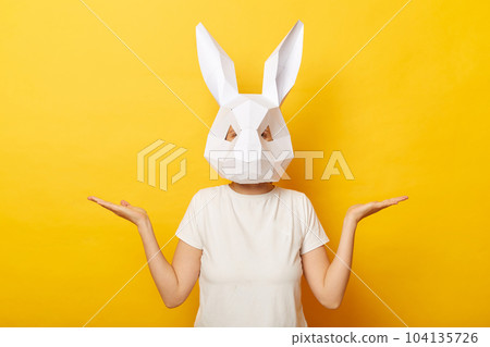 Horizontal shot of woman wearing white T-shirt and paper rabbit mask standing isolated over yellow background, posing with spread hands, presenting advertisement area on her palms. 104135726