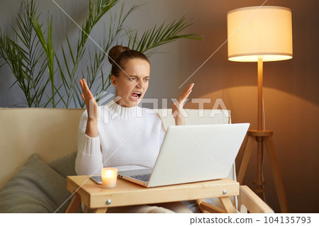 Portrait of aggressive angry woman wearing white sweater sitting on sofa and working on laptop in home interior, having trouble with her work, screaming with hate. Portrait of aggressive angry woman wearing white sweater sitting on sofa and working on laptop in home interior, having trouble with her work, screaming with hate. 104135793