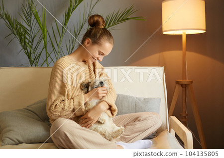 Young pretty woman in casual clothes hugging her beloved Pekingese dog sitting on the sofa in the living room of her cozy house, female smiling happily. 104135805