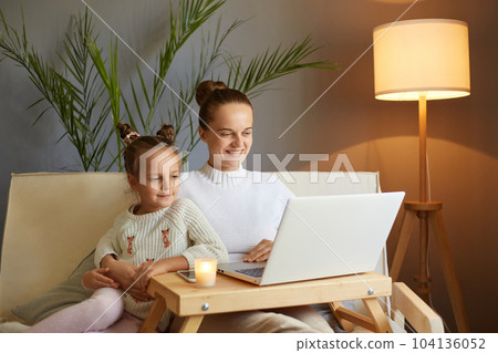 Horizontal shot of smiling woman with her cute little daughter, wearing white sweater sitting on sofa and watching videos or cartoons, spending time together. 104136052