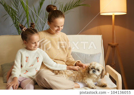 Indoor shot of mother and little daughter playing with dog on the home sofa, happy girl with Pekingese dog and mother relaxing at home, family with pet having fun together. 104136055