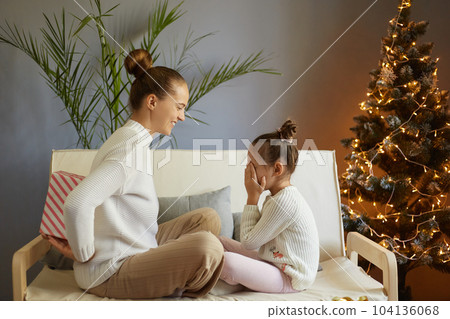 Happy family sharing joy with each other on Christmas. Daughter receives gifts from her loving mother on Xmas Day. Kid covering her eyes, waiting for surprise, sitting in decorated room. Happy family sharing joy with each other on Christmas. Daughter receives gifts from her loving mother on Xmas Day. Kid covering her eyes, waiting for surprise, sitting in decorated room. 104136068