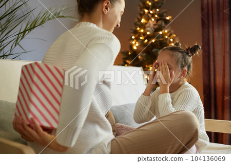 Indoor shot of woman wearing white jumper posing backwards giving present box to her daughter while sitting near Christmas tree, celebrating new year, kid covering eyes, waiting her gift. Indoor shot of woman wearing white jumper posing backwards giving present box to her daughter while sitting near Christmas tree, celebrating new year, kid covering eyes, waiting her gift. 104136069