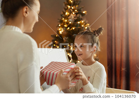 Back view of mother giving gift for her loving child on Xmas Day, daughter with mommy sitting on sofa in living room with decorated tree in background 104136070