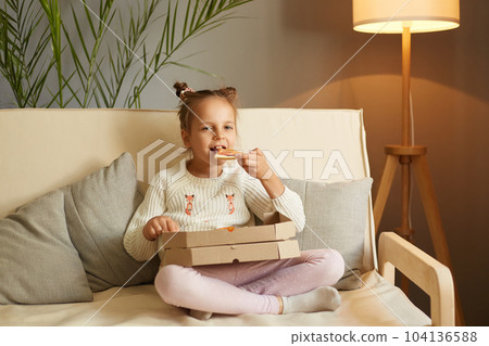Indoor shot of cute little girl sitting on couch and eating piece of italian Pizza at home, yummy meal, kid wearing casual clothing with funny hair buns enjoying delicious junk food. 104136588