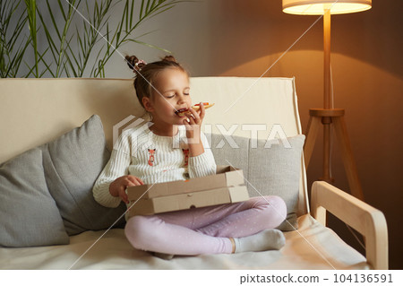 Full length portrait of hungry adorable female kid sitting on cough with crossed legs and eating fast food, holding slice of pizza and biting tasty snack. 104136591