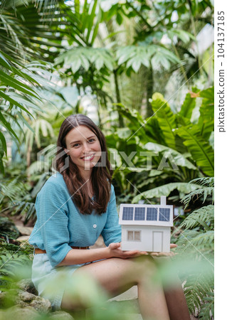 Young woman in jungle holding paper model of house with solar panels, concept of renewable energy and protection of nature. 104137185