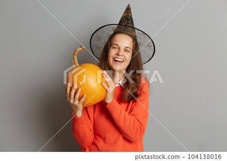 Image of extremely happy woman wearing witch hat standing with orange pumpkin, celebrating Halloween, laughing out loud, posing isolated over gray background. 104138016