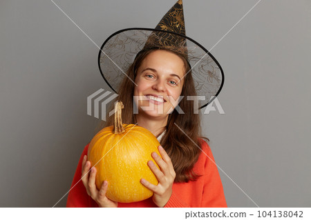 Horizontal shot of smiling optimistic attractive woman holding big ripe orange pumpkin, looking at camera with toothy smile, celebrating halloween, standing isolated over gray background. Horizontal shot of smiling optimistic attractive woman holding big ripe orange pumpkin, looking at camera with toothy smile, celebrating halloween, standing isolated over gray background. 104138042