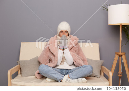 Portrait of frozen beautiful woman wearing warm clothes and wrapped in scarf sitting on sofa in room, trying to get warm in cold apartment, looking at camera. 104138050