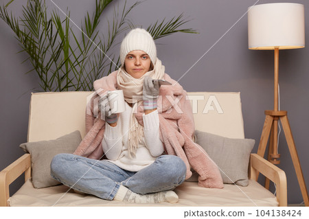 Portrait of winsome sad upset cold woman sitting on the sofa at home drinking hot tea and using smart phone, female wearing winter coat, hat and mittens. 104138424
