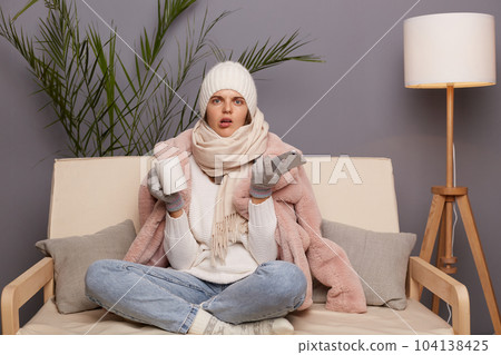 Portrait of confused young adult woman wearing warm clothes and wrapped in scarf sitting on cough with cup of hot tea, being confused, using smart phone. Portrait of confused young adult woman wearing warm clothes and wrapped in scarf sitting on cough with cup of hot tea, being confused, using smart phone. 104138425