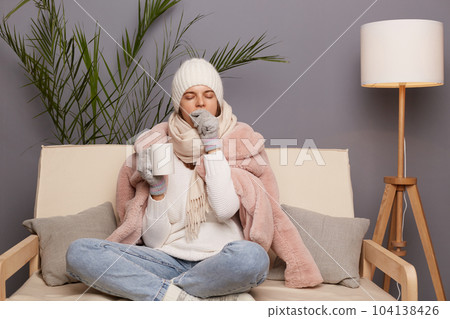 Indoor shot of sleepy Caucasian woman wearing warm clothes and wrapped in scarf sitting on sofa at home with cup of hot tea, being sleepless, covering mouth with hand while yawning. 104138426