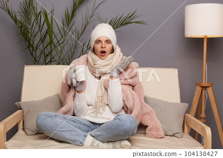Image of shocked amazed beautiful woman wearing warm clothes and wrapped in scarf sitting on cough in living room with cup of hot tea, keeps legs crossed, looking at camera with open mouth. 104138427