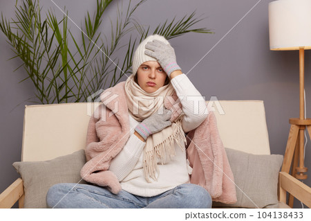 Indoor shot of sick unhealthy frozen woman wearing cap, gloves, scarf and coat sitting in living room, fells terrible headache and flu symptoms, very cold in flat in winter. 104138433
