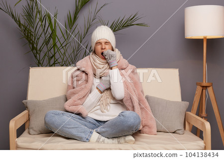 Full length portrait of sleepy woman posing in cold room sitting on the cough wearing warm hat, mittens and cap, needs to have nap, yawning, covering mouth with hand. 104138434
