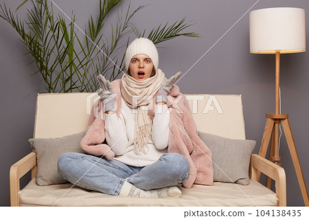 Shocked angry Caucasian woman sitting on the sofa at home, looking at camera with open mouth and big eyes, asking why central heating is turn off, female wearing winter coat, hat and mittens. 104138435