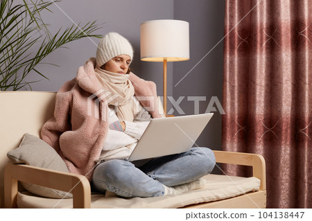 Side view portrait of calm sad depressed woman wearing cap and winter coat sitting on the sofa at home, holding laptop, looking at display with disappointed expressing, fells cold and bored. 104138447