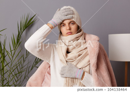 Image of ill attractive unhealthy young adult woman wearing cap and coat posing in living room, catching cold, having high temperature and terrible headache. Image of ill attractive unhealthy young adult woman wearing cap and coat posing in living room, catching cold, having high temperature and terrible headache. 104138464