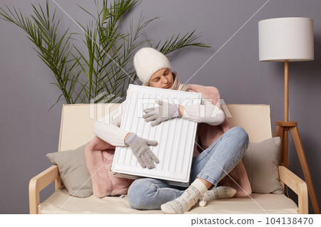 Indoor shot of sleepy frozen woman in coat and hat sit in cold living room and embracing radiator, feels freeze at home, having nap while getting warm, keeps eyes closed. Indoor shot of sleepy frozen woman in coat and hat sit in cold living room and embracing radiator, feels freeze at home, having nap while getting warm, keeps eyes closed. 104138470