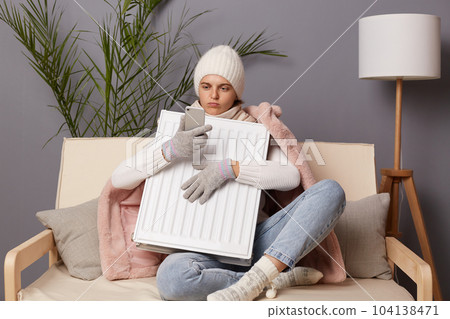 Portrait of Caucasian woman in coat and hat sit in cold living room embracing electric heating to get warm and using cell phone, call to service, broken central heating. Portrait of Caucasian woman in coat and hat sit in cold living room embracing electric heating to get warm and using cell phone, call to service, broken central heating. 104138471