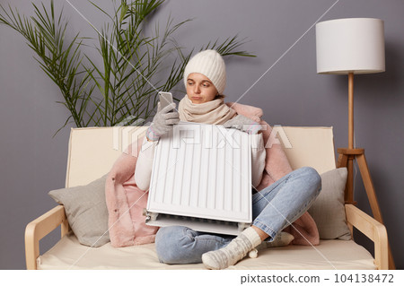 Horizontal shot of confused woman wearing cap and winter coat sitting on the sofa at home, embracing battery, trying to get warm, calling to service, problems with heating. 104138472