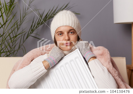 Portrait of sad disappointed young adult woman in coat and hat sit in cold living room with heater in hands, looking at camera with upset expression and pout lips. Portrait of sad disappointed young adult woman in coat and hat sit in cold living room with heater in hands, looking at camera with upset expression and pout lips. 104138476