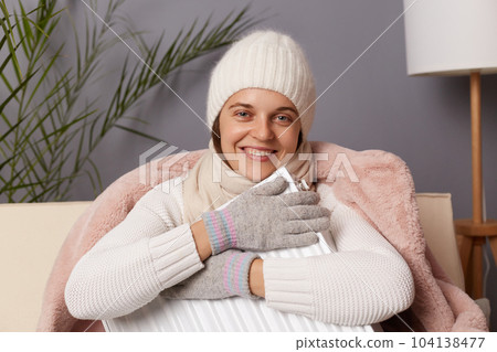 Image of smiling delighted woman in coat and hat sit in cold living room with radiator, looking at camera, embracing heater, being in good mood despite the lack of heat in her apartment. Image of smiling delighted woman in coat and hat sit in cold living room with radiator, looking at camera, embracing heater, being in good mood despite the lack of heat in her apartment. 104138477