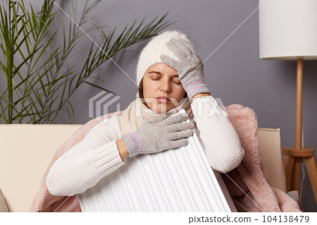 Horizontal shot of sick unhealthy ill Caucasian woman wearing warm cap and gloves embracing battery, posing in home interior, keeps hand on painful headache, sitting with closed eyes.. 104138479