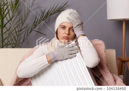 Portrait of sick unhealthy woman in coat and hat sit in cold living room with radiator, has no heating in her house, suffering flu and headache, looking at camera with sad expression. 104138480