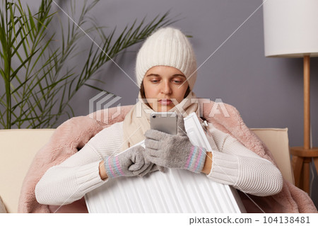 Image of attractive young adult serious concentrated woman wearing warm cap and gloves embracing heater, posing in home interior, using cell phone, search information about central heating service. 104138481
