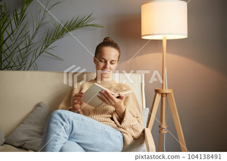 Indoor shot of pleased pretty brown haired woman wearing beige jumper and jeans sitting on cough with book in hands, reading interesting novel or beautiful poems. Indoor shot of pleased pretty brown haired woman wearing beige jumper and jeans sitting on cough with book in hands, reading interesting novel or beautiful poems. 104138491