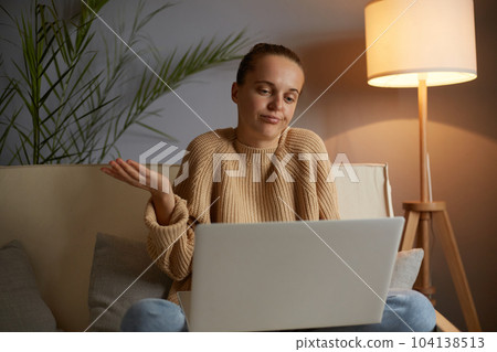 Indoor shot of puzzled confused Caucasian woman wearing beige jumper and jeans sitting on cough in lotus pose, having video call on laptop, shrugging shoulders, looks helpless. 104138513