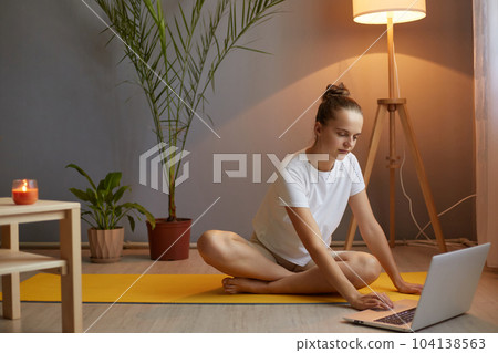 Portrait of concentrated young adult woman with hair bun wearing white t shirt practicing yoga, using laptop and internet for choosing online course. 104138563