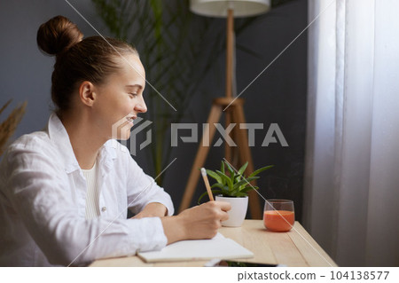 Image of smiling positive optimistic young beautiful woman writing down notes while sitting at table in office, thinking about to do list, expressing positive emotions. 104138577