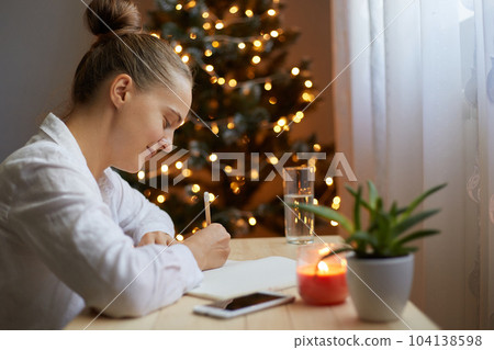 Smiling joyful woman in white shirt sitting at table and writing Christmas bucket list or tasks she needs to do in preparation for winter holidays, writes in a notebook in room with xmas tree. 104138598