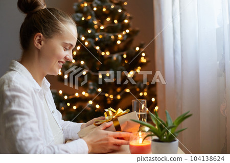 Profile portrait of smiling beautiful woman wearing white shirt sitting on table near christmas tree and holding present box in hands, being very happy, waiting for holidays. Profile portrait of smiling beautiful woman wearing white shirt sitting on table near christmas tree and holding present box in hands, being very happy, waiting for holidays. 104138624